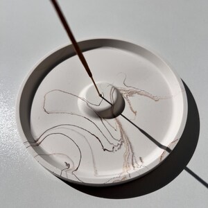 May include: A round, white incense holder with brown and tan marbling. The holder has a central hole for the incense stick, which is lit and emitting smoke. The design is minimalist and modern, suitable for home decor.