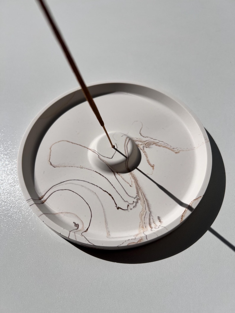 May include: A round, white incense holder with brown and tan marbling. The holder has a central hole for the incense stick, which is lit and emitting smoke. The design is minimalist and modern, suitable for home decor.