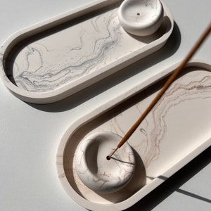 Könnte beinhalten: Zwei weiße ovale Keramikschalen für Räucherstäbchen mit Marmormuster. Eine Schale hat ein brennendes braunes Räucherstäbchen in der Mitte.