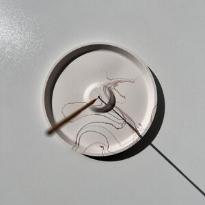 May include: A round, white incense holder with a dark rim and abstract brown and black ink patterns. An incense stick is placed in the center, casting a shadow on the white surface. The holder is on a light gray surface.