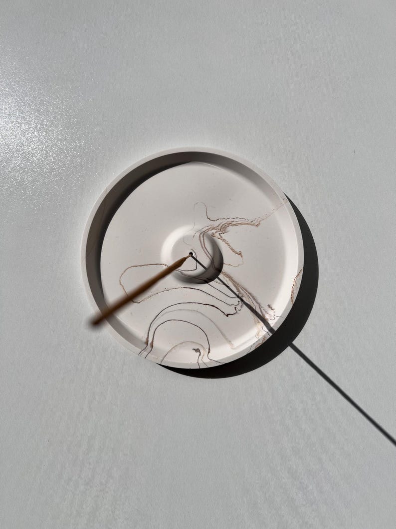 May include: A round, white incense holder with a dark rim and abstract brown and black ink patterns. An incense stick is placed in the center, casting a shadow on the white surface. The holder is on a light gray surface.
