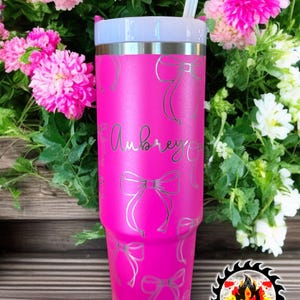 May include: A bright pink tumbler with a silver lid and straw, adorned with silver bow designs. The name "Aubrey" is scripted on the front. The tumbler is surrounded by pink and white flowers.