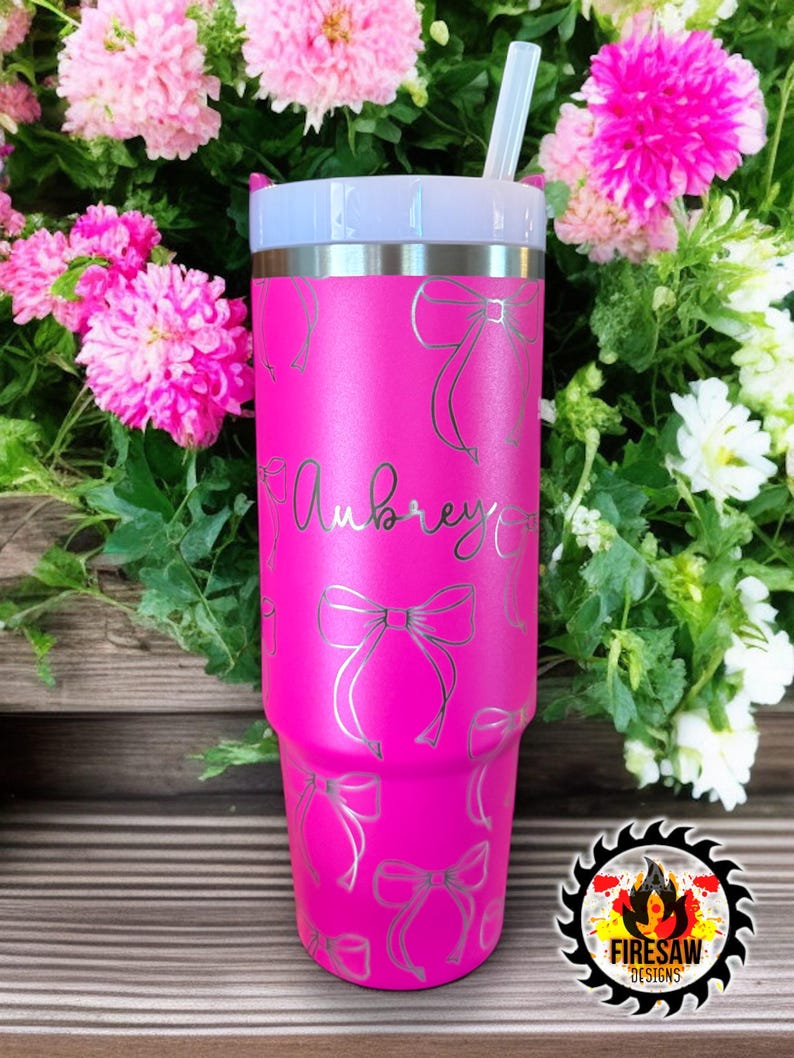 May include: A bright pink tumbler with a silver lid and straw, adorned with silver bow designs. The name "Aubrey" is scripted on the front. The tumbler is surrounded by pink and white flowers.