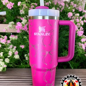 May include: A bright pink Stanley tumbler with a handle and a white lid. The tumbler features a pattern of silver bows and the Stanley logo. A white straw is inserted into the lid.