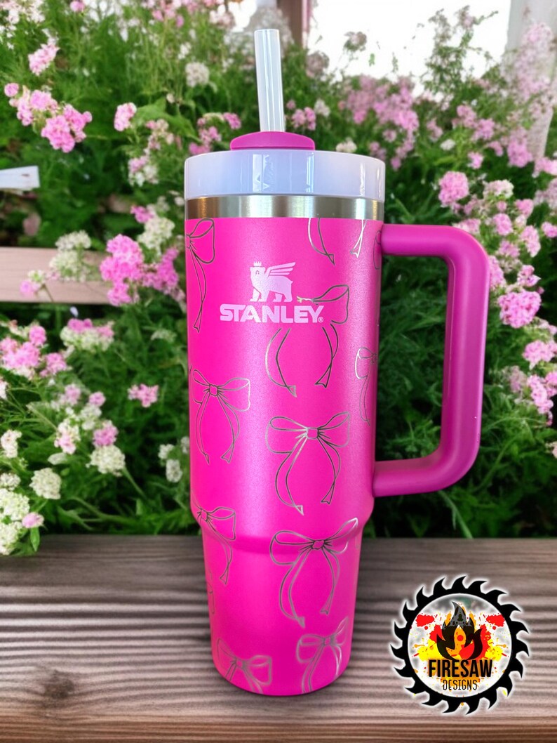 May include: A bright pink Stanley tumbler with a handle and a white lid. The tumbler features a pattern of silver bows and the Stanley logo. A white straw is inserted into the lid.