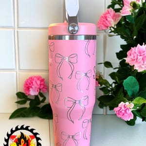May include: A pink insulated tumbler with a silver bow pattern. The tumbler has a white handle and a silver rim. The design includes the text "Firesaw Designs" on the bottom left. Pink flowers are in the background.