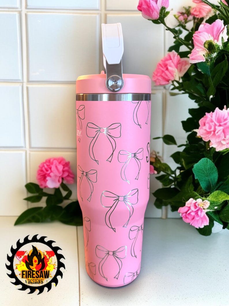 May include: A pink insulated tumbler with a silver bow pattern. The tumbler has a white handle and a silver rim. The design includes the text "Firesaw Designs" on the bottom left. Pink flowers are in the background.