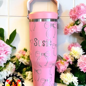 May include: A pink insulated tumbler with a white handle and lid. The tumbler is decorated with silver bow designs and the name "Susie". The tumbler is surrounded by pink flowers.
