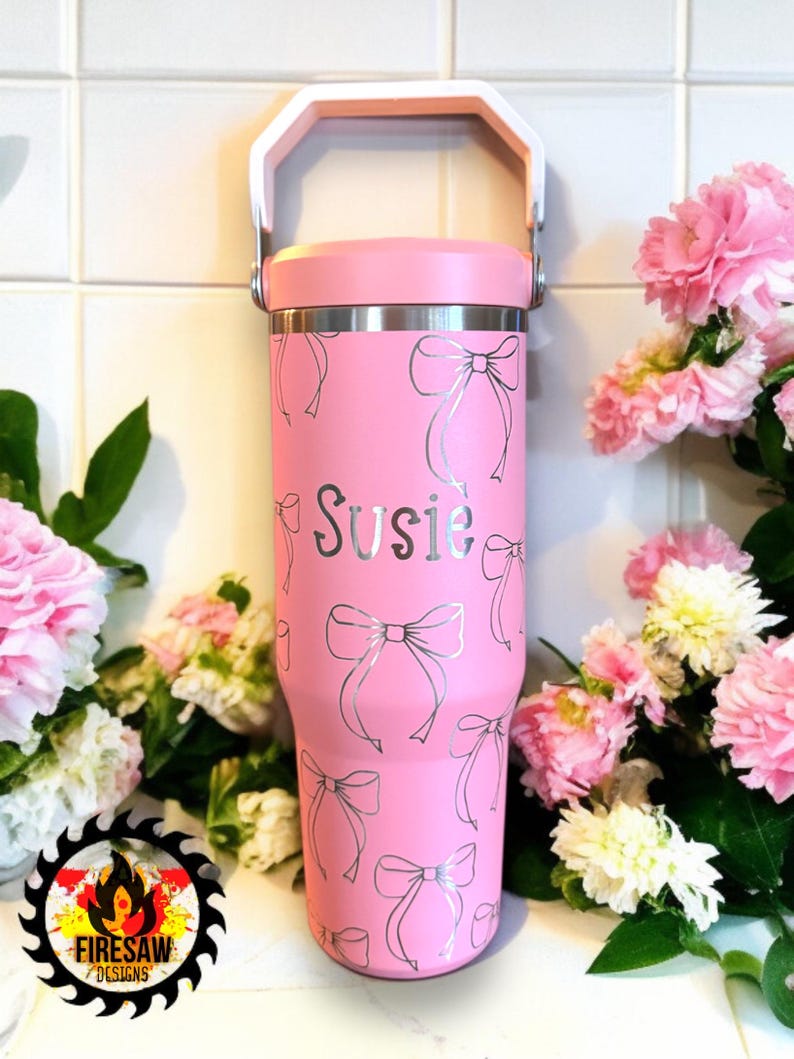 May include: A pink insulated tumbler with a white handle and lid. The tumbler is decorated with silver bow designs and the name "Susie". The tumbler is surrounded by pink flowers.