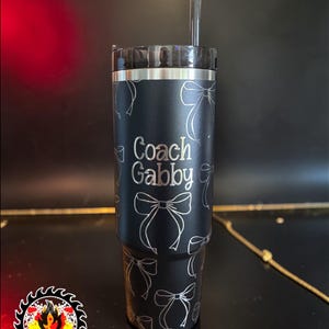 May include: Black tumbler with a silver lid and straw. The tumbler is decorated with a pattern of silver bows and the words "Coach Gabby". The Firesaw Designs logo is in the lower left corner.