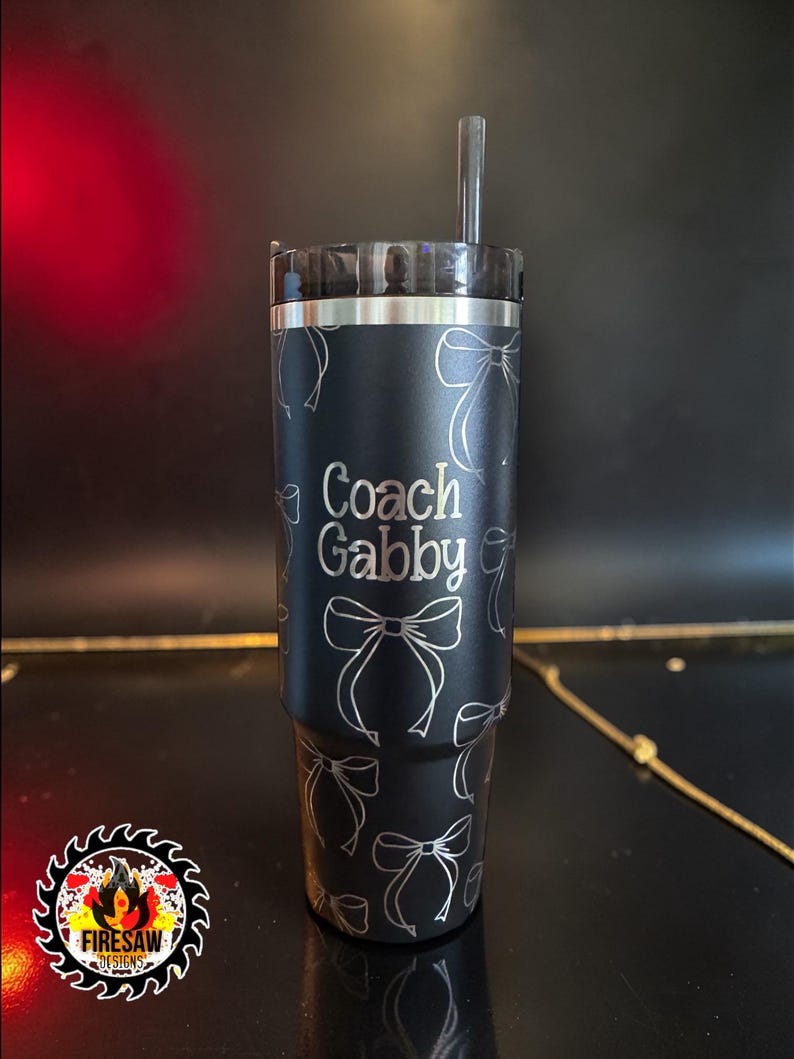 May include: Black tumbler with a silver lid and straw. The tumbler is decorated with a pattern of silver bows and the words "Coach Gabby". The Firesaw Designs logo is in the lower left corner.