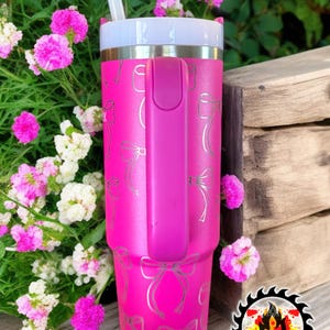 May include: A bright pink tumbler with a silver bow pattern and a matching pink handle. The tumbler has a white lid and a white straw. The tumbler is next to pink and white flowers.