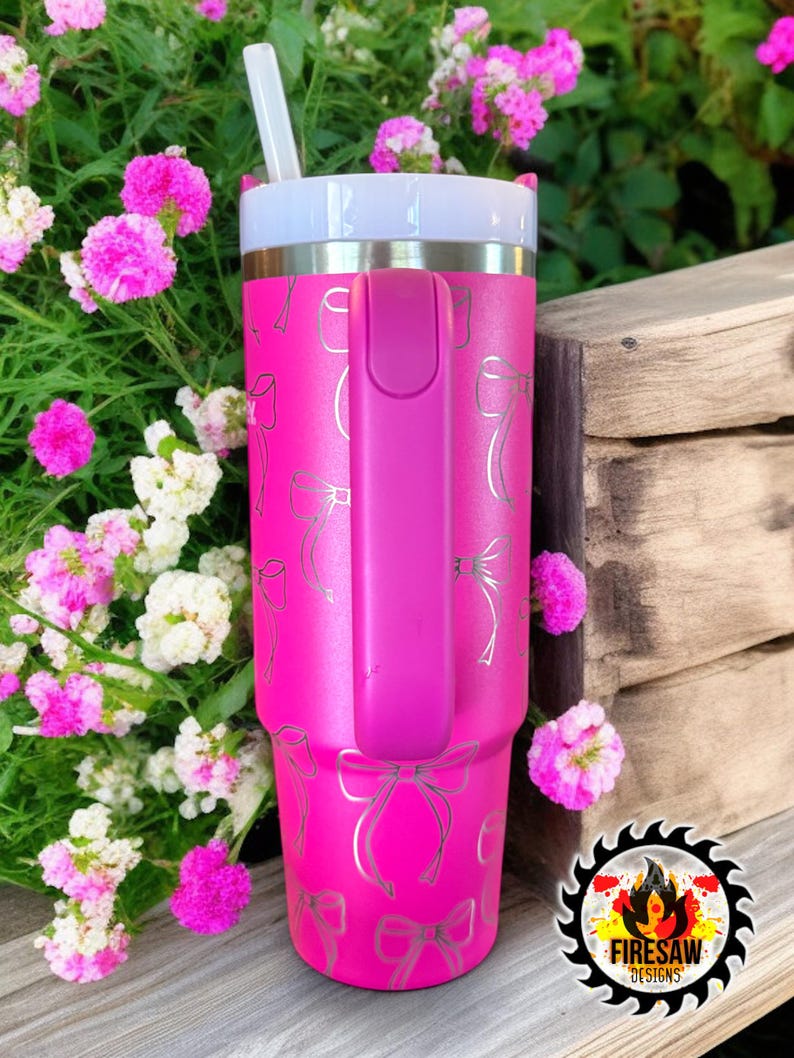 May include: A bright pink tumbler with a silver bow pattern and a matching pink handle. The tumbler has a white lid and a white straw. The tumbler is next to pink and white flowers.