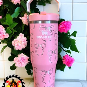 May include: A pink Stanley tumbler with a white handle and lid. The tumbler is decorated with a pattern of silver bows. The Stanley logo is visible on the front. Pink flowers and green leaves are in the background.