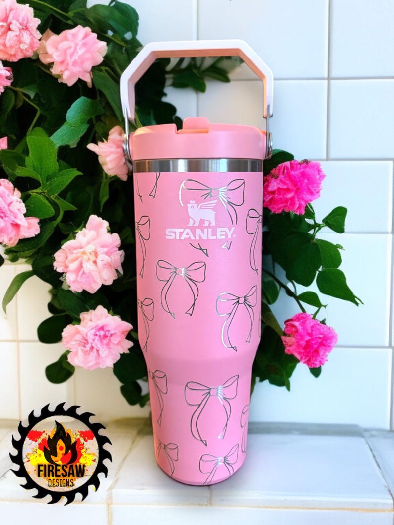 May include: A pink Stanley tumbler with a white handle and lid. The tumbler is decorated with a pattern of silver bows. The Stanley logo is visible on the front. Pink flowers and green leaves are in the background.