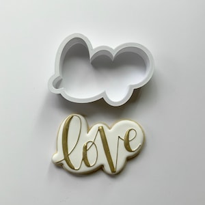 Op de afbeelding: Een witte koekjesvormer in de vorm van een wolk met het woord "love" geschreven in gouden glazuur op een witte koek.