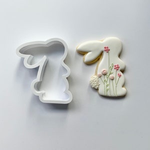 May include: White cookie cutter and a decorated cookie in the shape of a bunny with pink flowers and green stems.
