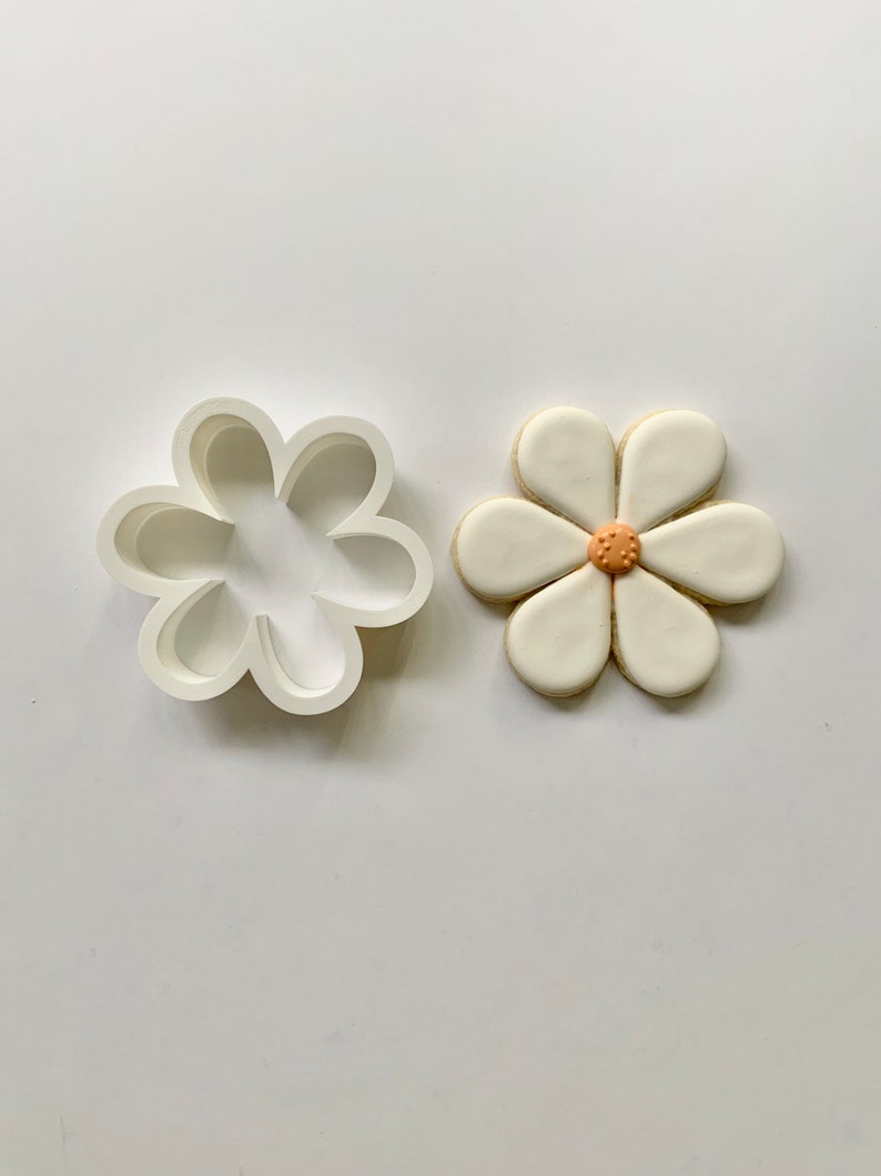 May include: A white cookie cutter shaped like a flower with six petals. Next to it is a white cookie decorated with a light orange center.