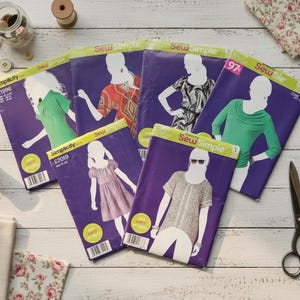 May include: A collection of Simplicity sewing patterns with various designs, including dresses and tops, arranged on a white wooden surface. The patterns are purple with illustrations of the garments. Accessories like buttons, scissors, and spools of thread are also present.