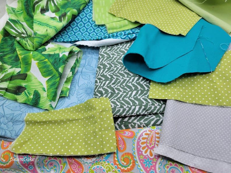 Op de afbeelding: Een assortiment van stoffenresten in verschillende patronen en kleuren, waaronder groen, blauw, wit en roze. De stoffen zijn in een stapel gerangschikt, waarbij sommige elkaar overlappen.