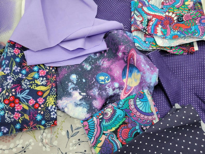Op de afbeelding: Een verzameling stofresten in verschillende kleuren en patronen, waaronder paars, blauw, roze en wit. De stoffen hebben bloemen-, galaxy- en paisley-ontwerpen.