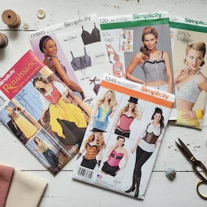 May include: A collection of Simplicity sewing patterns featuring vintage and costume designs. The patterns showcase various styles, including Renaissance and 1950s vintage looks. Sewing notions like spools of thread, buttons, and scissors are also visible.