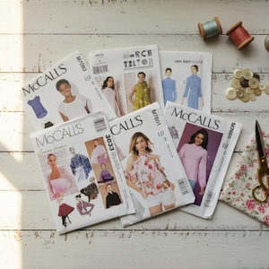 May include: A collection of sewing patterns from McCall's and other brands, featuring various garment designs. The image includes spools of thread, buttons, scissors, and floral fabric, suggesting a sewing or crafting theme.