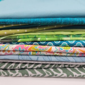 Op de afbeelding: Een stapel stofresten in verschillende tinten blauw, groen en roze. De stof heeft verschillende patronen, waaronder bloemen, paisley en geometrische ontwerpen.