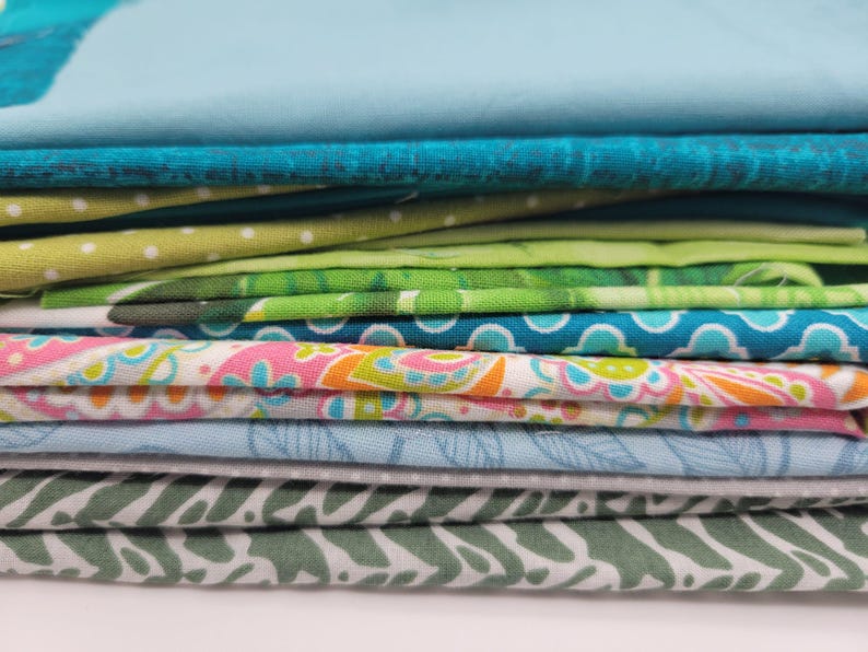 Op de afbeelding: Een stapel stofresten in verschillende tinten blauw, groen en roze. De stof heeft verschillende patronen, waaronder bloemen, paisley en geometrische ontwerpen.