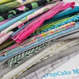 Op de afbeelding: Een stapel kleurrijke stoffenresten in verschillende patronen en kleuren, waaronder roze, blauw, groen en wit. De stoffenresten zijn in een stapel gerangschikt, met de bovenkant van de stapel die een verscheidenheid aan patronen laat zien. De stoffenresten zijn gelabeld met "ScrapCake Mix" en "Sew happy!"