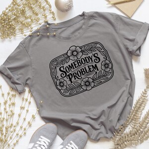 Puede incluir: Una camiseta gris con el texto "SOMEBODY'S PROBLEM" dentro de un marco floral en blanco y negro. La camiseta está colocada plana, con un par de zapatillas grises en la parte inferior.