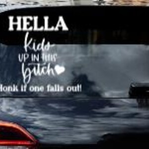 Op de afbeelding: Een witte vinyl sticker op de achterruit van een auto. De tekst luidt "HELLA kids UP IN THIS bitch" met een hartsymbool en "Toeter als er een uitvalt!"