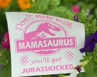 May include: A white rectangular decal featuring a pink graphic of a dinosaur silhouette inside a circle. The text reads Don't mess with MAMASAURUS you'll get JURASSKICKED in a pink font, accompanied by three claw marks on each side of the bottom text.