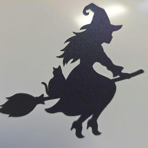 Op de afbeelding: Een zwarte wanddecoratie in silhouetvorm met een heks die een puntmuts en een jurk draagt en op een bezemsteel rijdt. Een kat zit op de bezemsteel achter de heks. Het ontwerp is plat en heeft een oppervlak met een textuur.