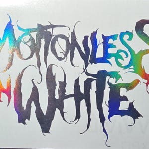 Op de afbeelding: Een holografische sticker met de tekst Motionless In White in een grillig, gestileerd lettertype. De letters vertonen een wisselend spectrum van iriserende kleuren, waaronder blauw, paars, groen en oranje op een witte achtergrond.