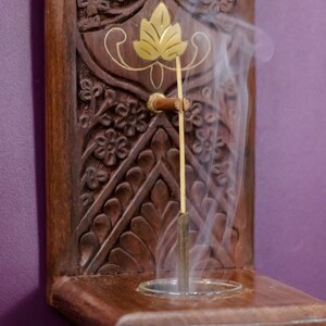 May include: A wooden incense holder with intricate carvings and a gold leaf design. A smoldering incense stick is burning in the holder, releasing a plume of white smoke.