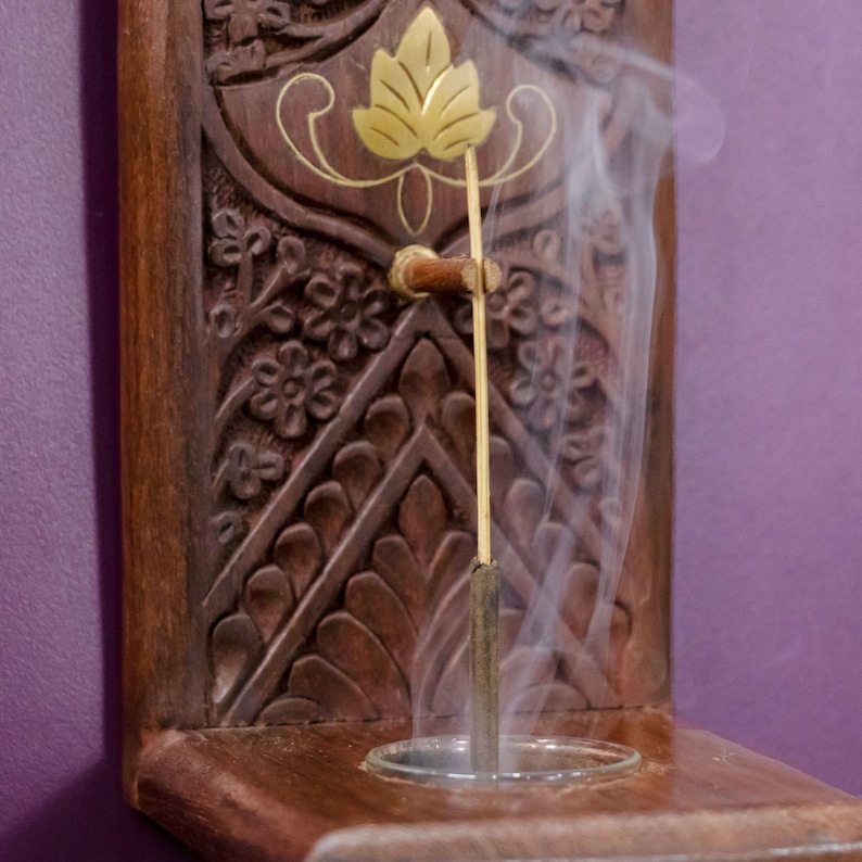 May include: A wooden incense holder with intricate carvings and a gold leaf design. A smoldering incense stick is burning in the holder, releasing a plume of white smoke.