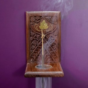 May include: A wooden incense holder with intricate carvings and a gold leaf design. A single incense stick is burning, releasing smoke.