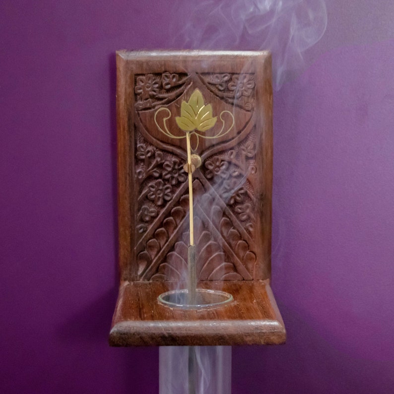May include: A wooden incense holder with intricate carvings and a gold leaf design. A single incense stick is burning, releasing smoke.