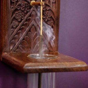 May include: A wooden incense holder with intricate carvings and a glass tube. A stick of incense is burning inside the tube, producing a plume of white smoke.