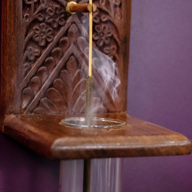 May include: A wooden incense holder with intricate carvings and a glass tube. A stick of incense is burning inside the tube, producing a plume of white smoke.