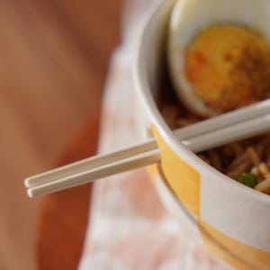 May include: A white bowl with a yellow rim filled with noodles and a hard-boiled egg.  Chopsticks are resting on the rim of the bowl.