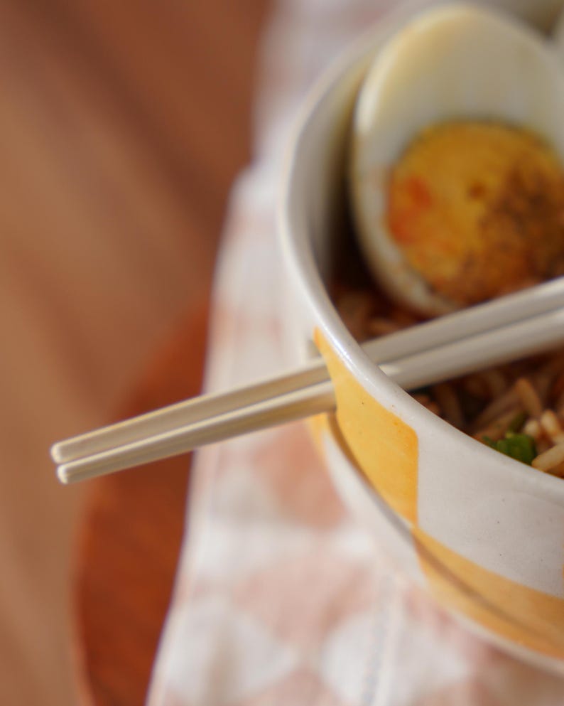 May include: A white bowl with a yellow rim filled with noodles and a hard-boiled egg.  Chopsticks are resting on the rim of the bowl.