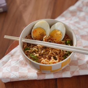 May include: A white bowl with yellow squares filled with noodles, green onions, and two halved hard-boiled eggs. White chopsticks are resting on the rim of the bowl.