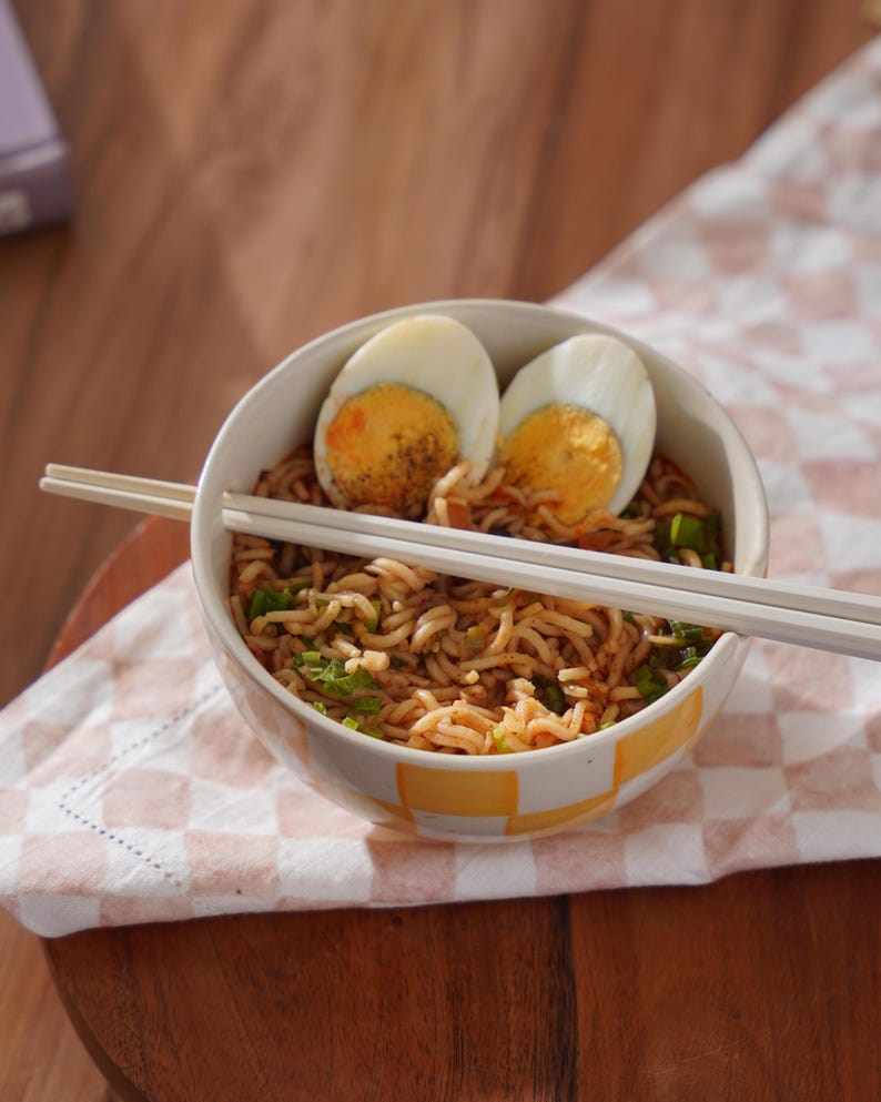 May include: A white bowl with yellow squares filled with noodles, green onions, and two halved hard-boiled eggs. White chopsticks are resting on the rim of the bowl.