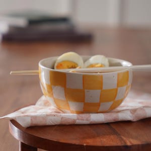 May include: A yellow and white checkered ceramic bowl with two halved hard-boiled eggs inside. The bowl is sitting on a wooden coaster with a white and pink checkered cloth napkin.