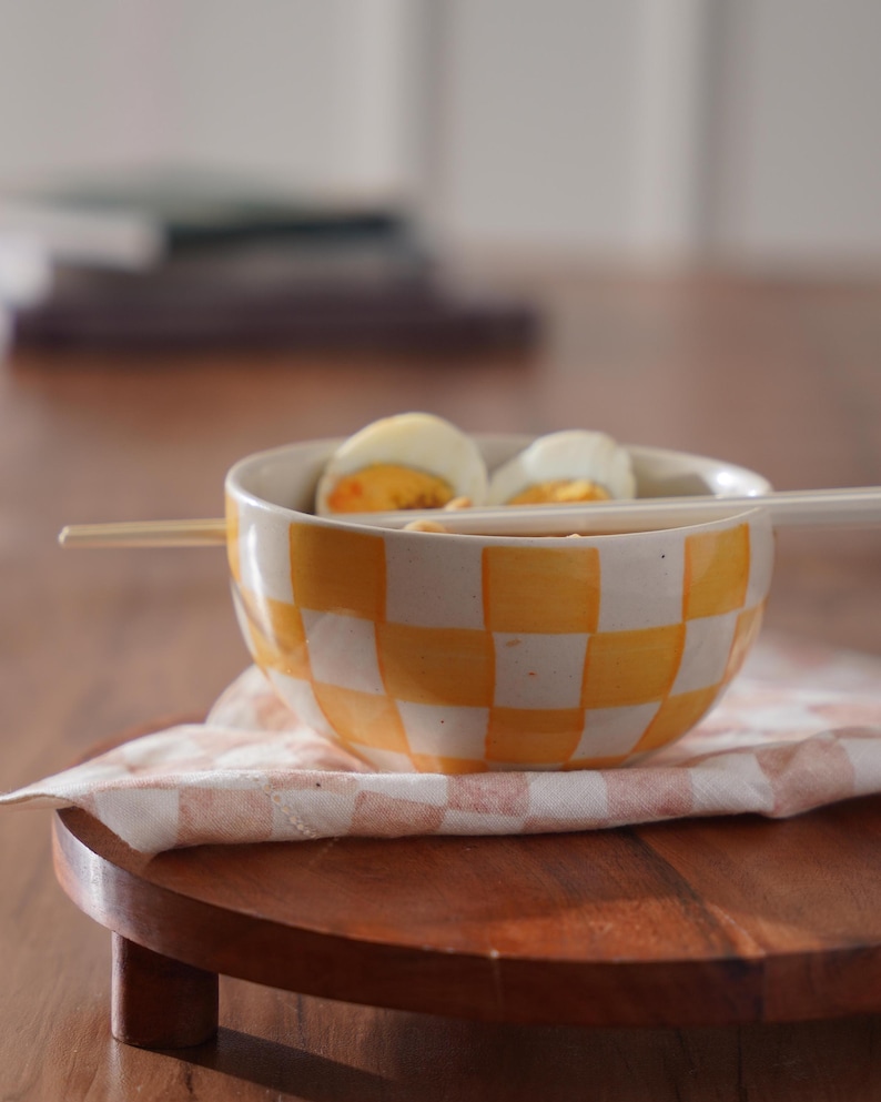 May include: A yellow and white checkered ceramic bowl with two halved hard-boiled eggs inside. The bowl is sitting on a wooden coaster with a white and pink checkered cloth napkin.