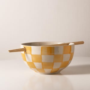 以下が含まれることがあります： 黄色と白の市松模様のセラミックボウルに、木製の箸が置かれている