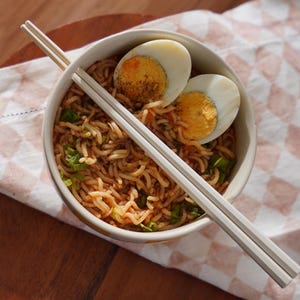 May include: A white bowl filled with noodles, chopped green onions, and two halved hard-boiled eggs. White chopsticks are laying across the top of the bowl.