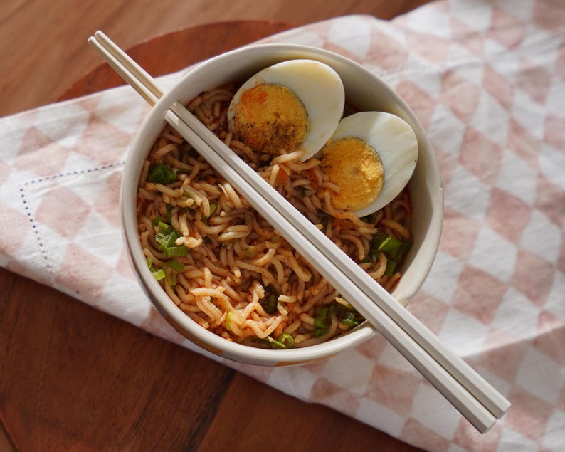 May include: A white bowl filled with noodles, chopped green onions, and two halved hard-boiled eggs. White chopsticks are laying across the top of the bowl.